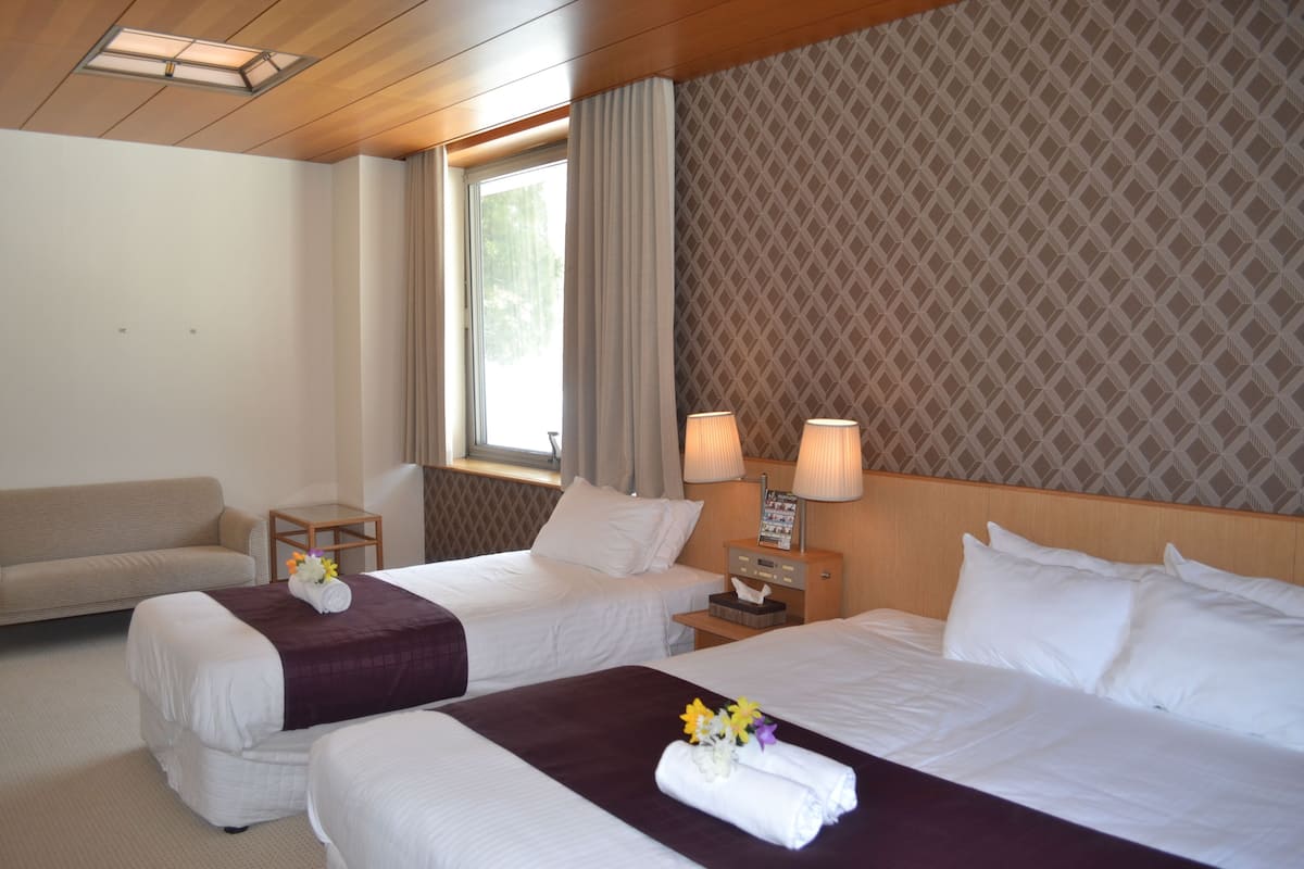 A hotel room with two beds, a sofa, a nightstand with lamps, and a window with curtains.