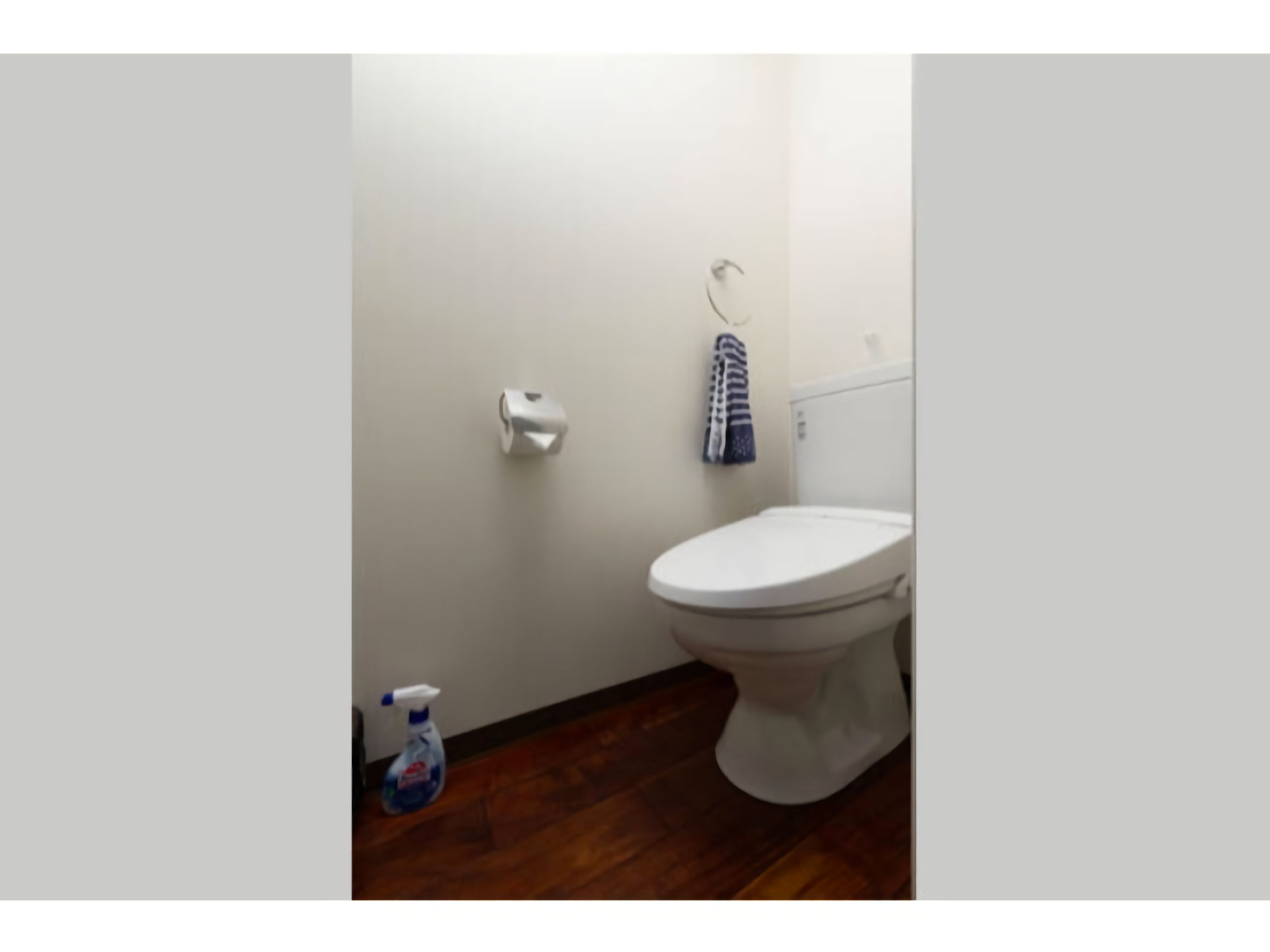 Une salle de bain avec des toilettes blanches, une serviette accrochée au mur et une bouteille de produit nettoyant sur le sol.