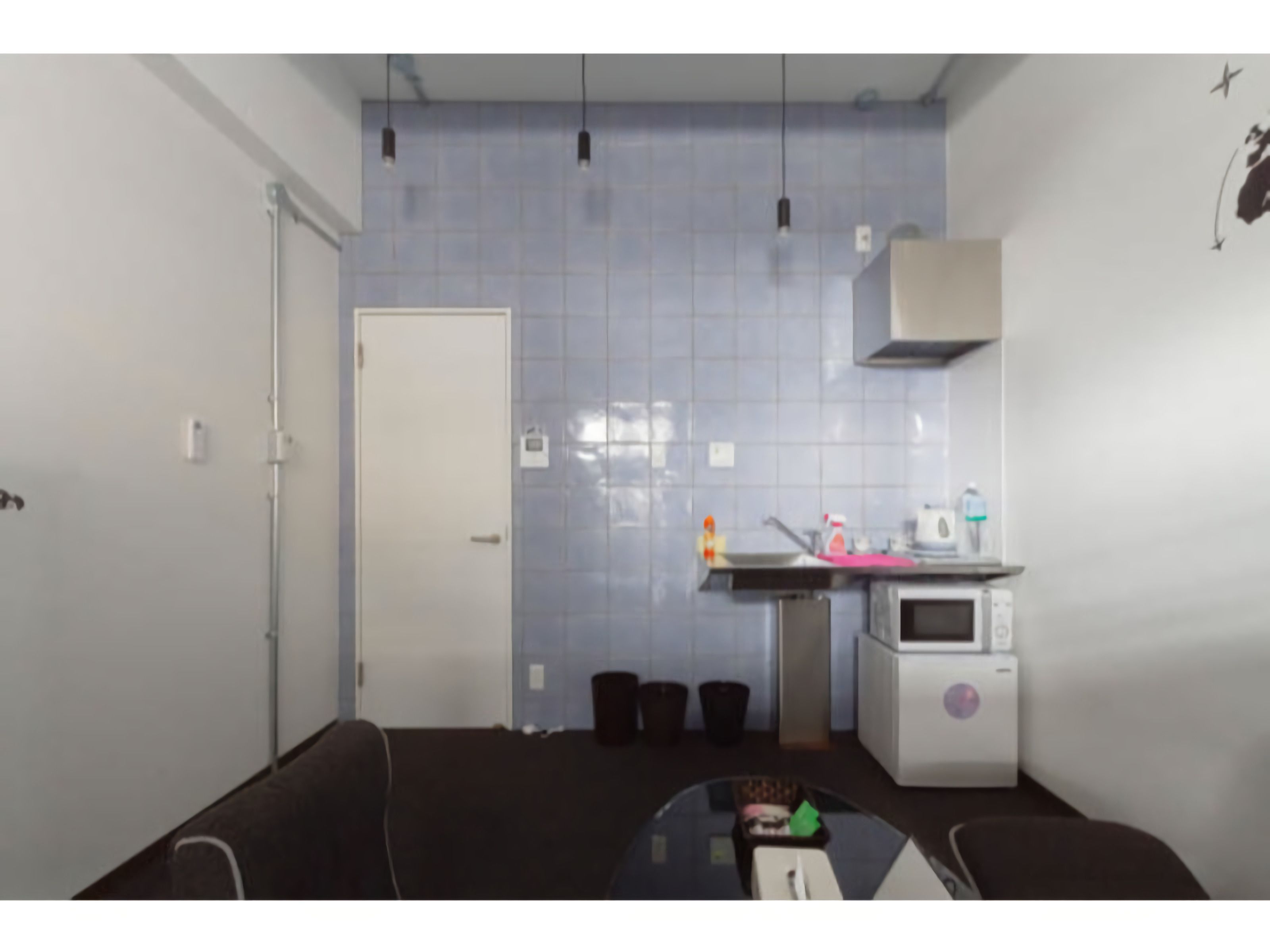 Cocina pequeña y funcional con microondas, lavamanos y armarios. Se observa una puerta que da a otra habitación y un cubo de basura cerca del lavamanos.