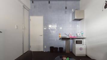 Cocina pequeña y funcional con microondas, lavamanos y armarios. Se observa una puerta que da a otra habitación y un cubo de basura cerca del lavamanos.
