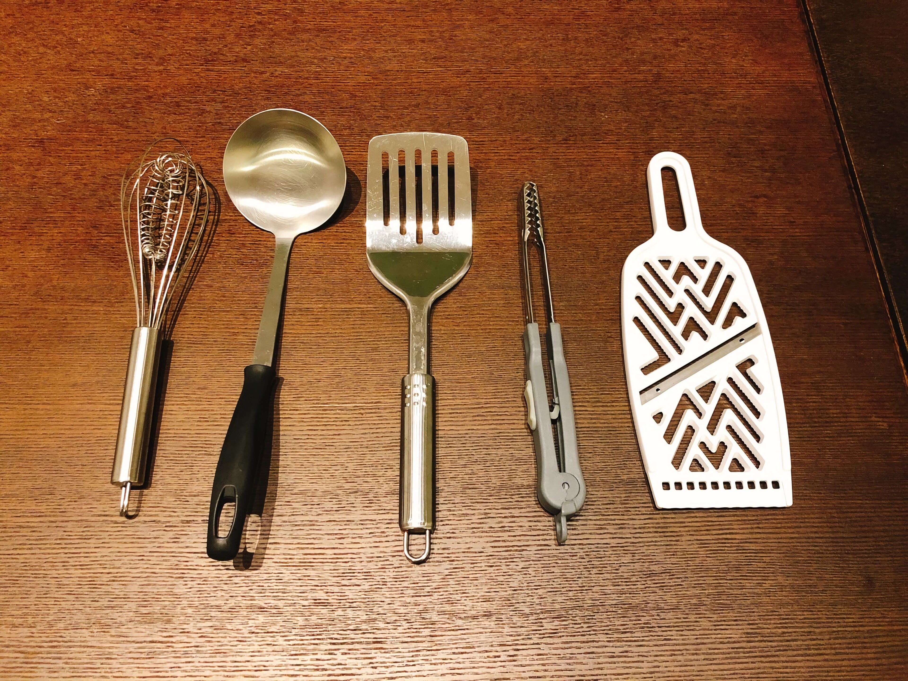 Superficie de madera con cinco utensilios de cocina: un batidor, una espumadera, una espátula, un par de pinzas y un rallador.