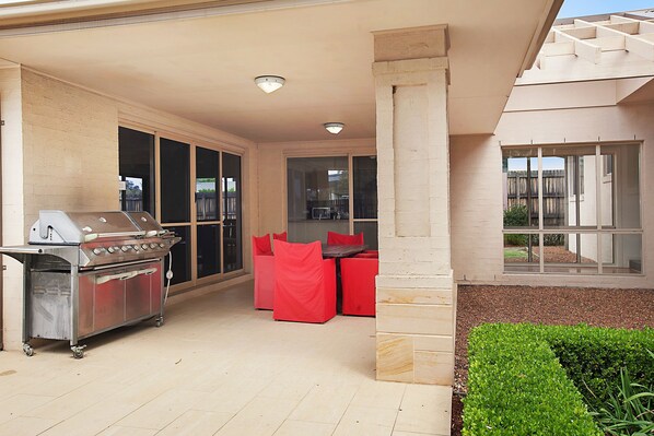 An outdoor patio with a grill, red chairs, and a view of the backyard. - Peppertree at the Vintage (Pokolbin)
