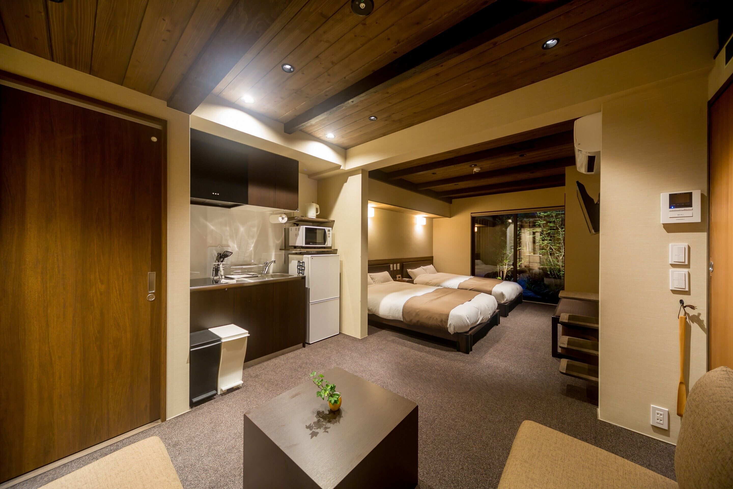 A hotel room with a kitchenette, a bed, a small table with a plant, and a view of the outside.