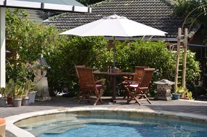 Een zwembadterras met een tafel en stoelen onder een grote parasol, omringd door weelderige begroeiing en potplanten.