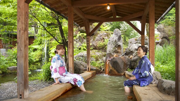 Two individuals sitting on wooden benches in a wooden structure with a water feature in the background. - Hirayuonsen Yamatokan (Takayama)