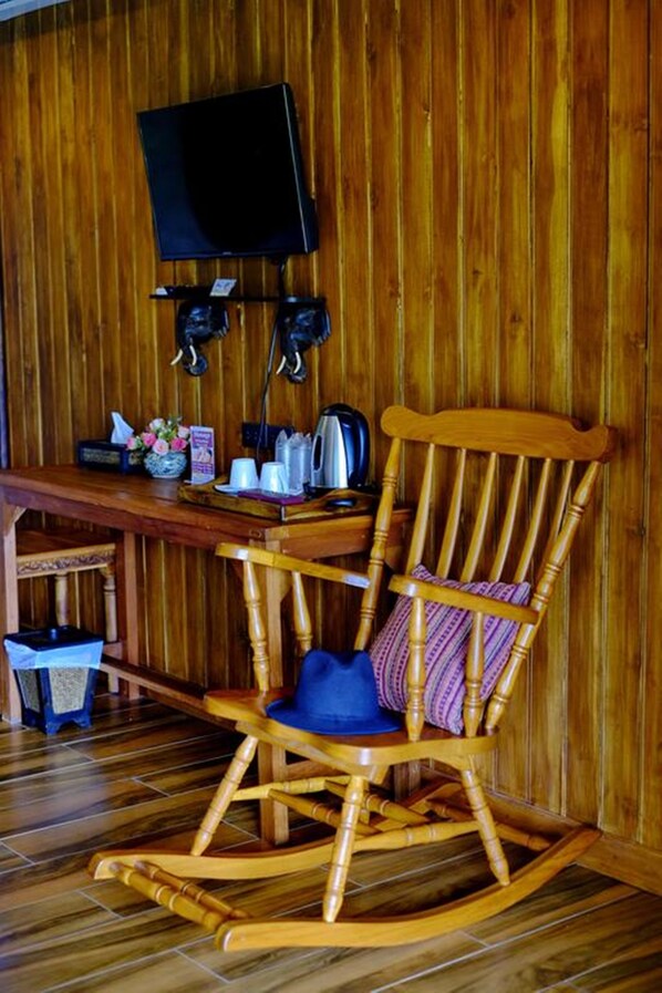 Una mecedora de madera con un cojín azul, una mesa de madera con un jarrón de flores y un televisor montado en la pared.
