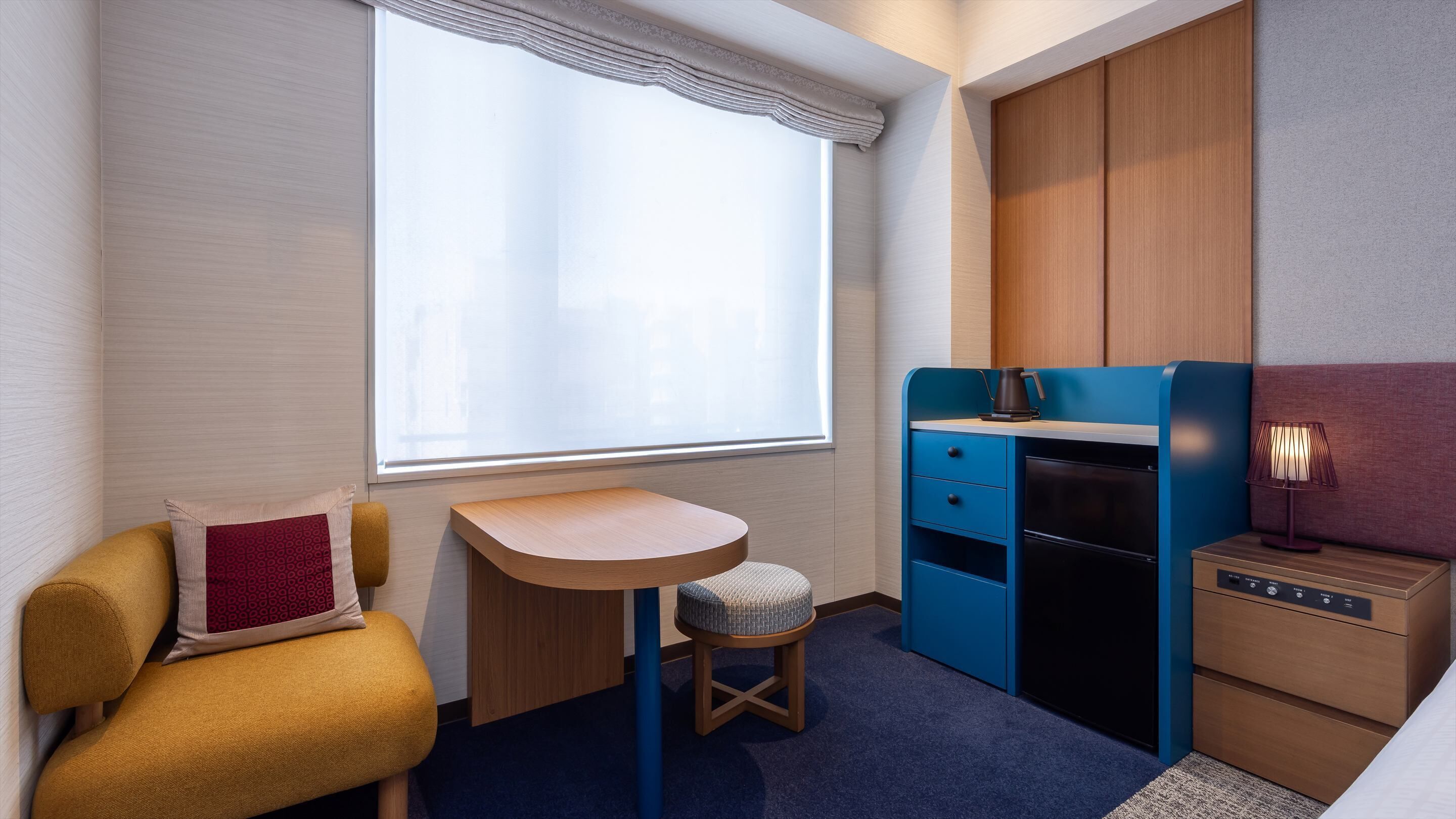 A compact room with a window, a small stool, a desk, a wardrobe, and a bedside table with a lamp.