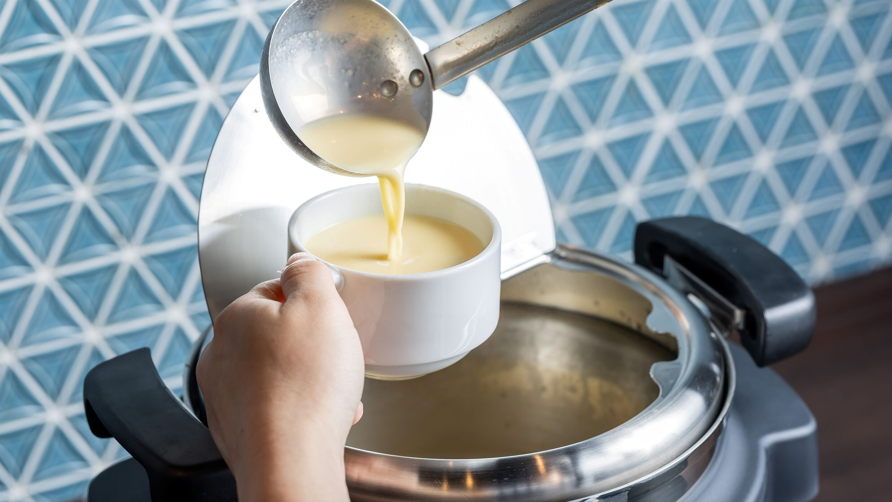 A hand pouring a creamy liquid from a metal container into a white cup.