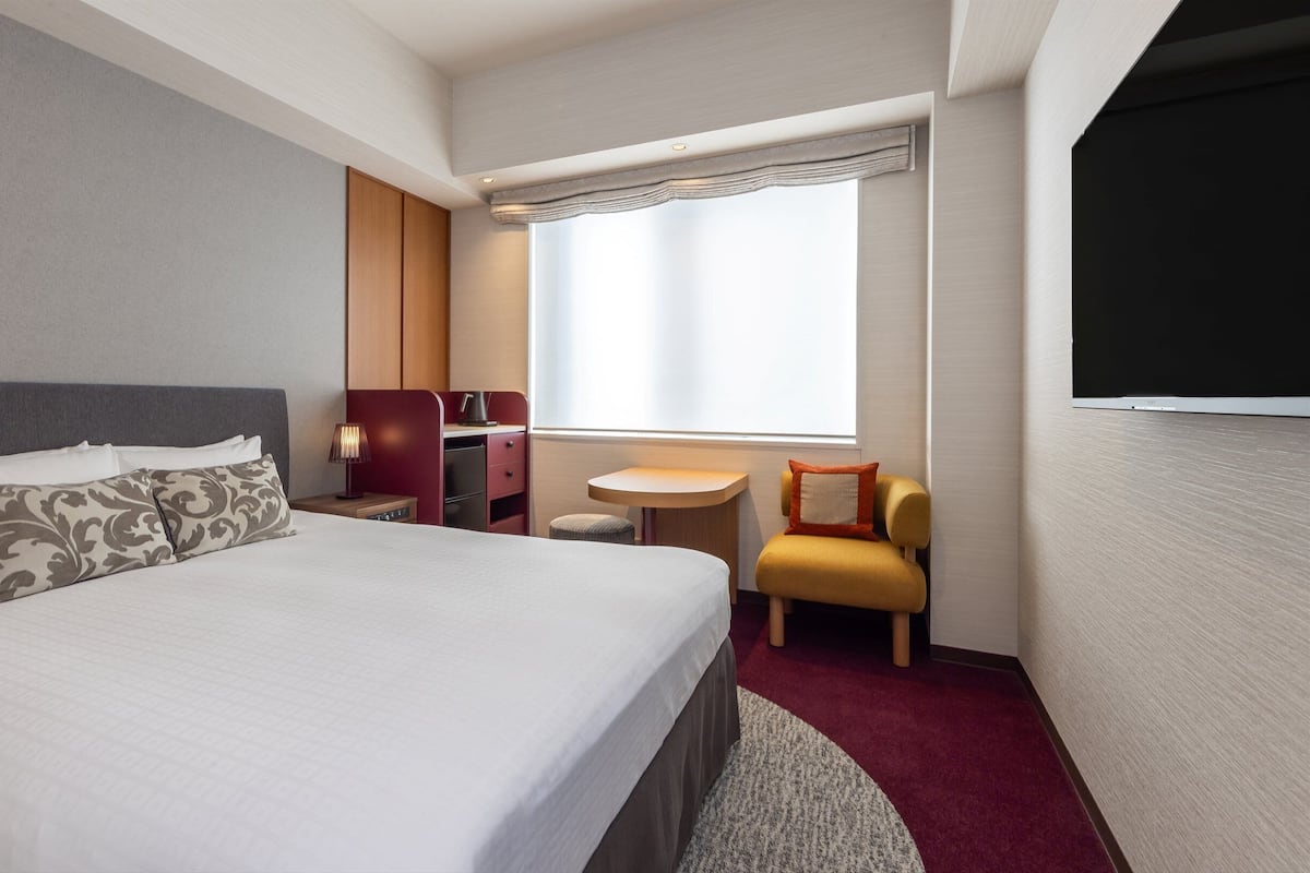 Une chambre d’hôtel avec un lit, un bureau, une chaise, une télévision et une fenêtre avec des rideaux.