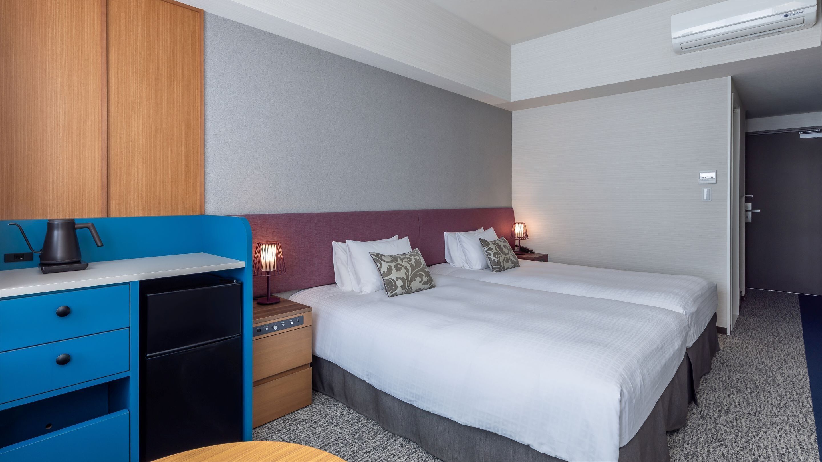Una habitación de hotel con cama, mesitas de noche, un escritorio con cajones, una lámpara y un equipo de aire acondicionado.