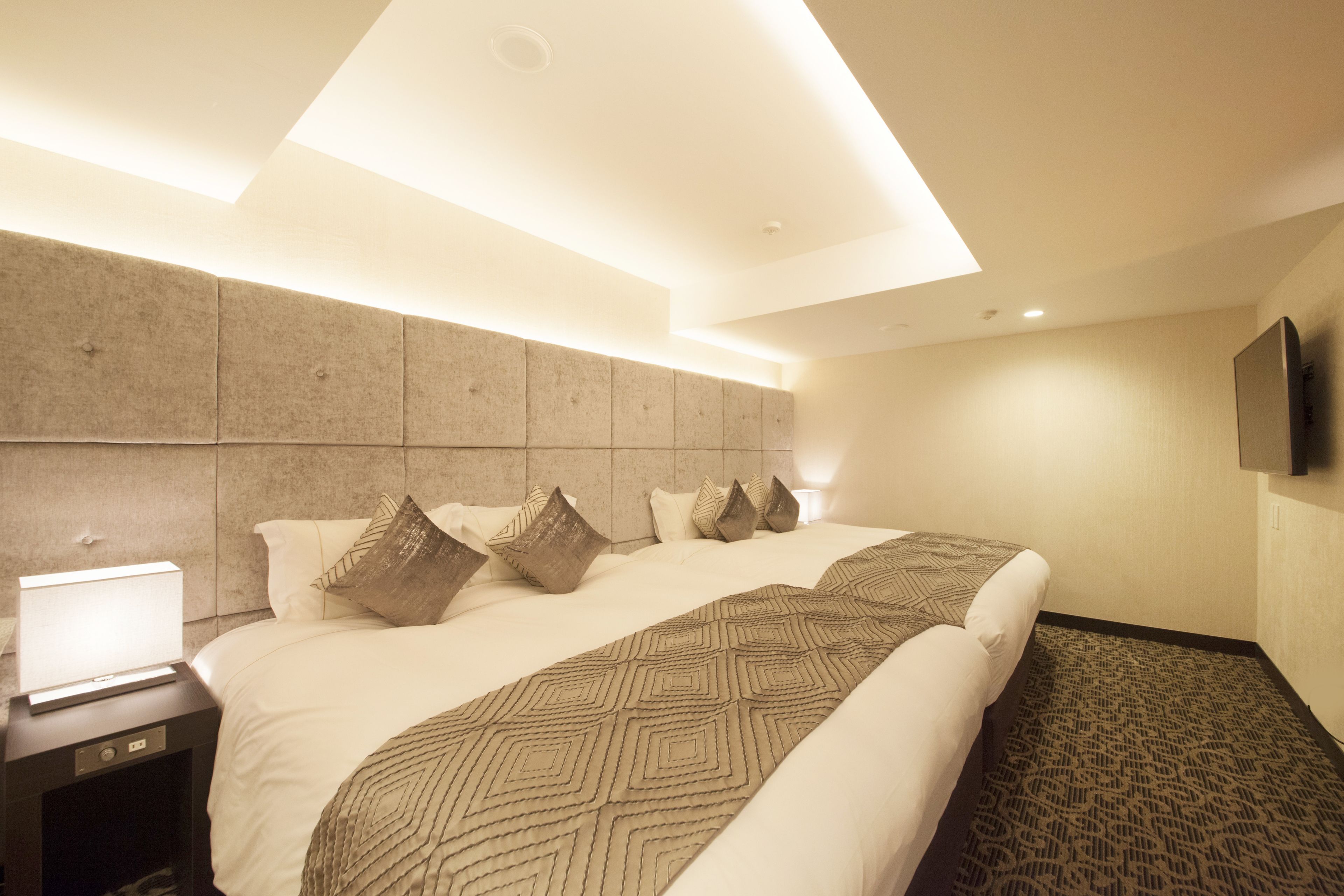 A modern hotel room with a large bed, a nightstand with a lamp, and a flat-screen TV mounted on the wall.