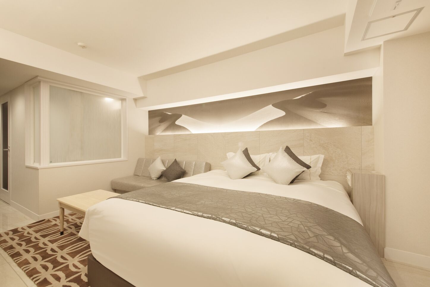 A modern bedroom with a large bed, a bedside table, a built-in bench, and a window with blinds.