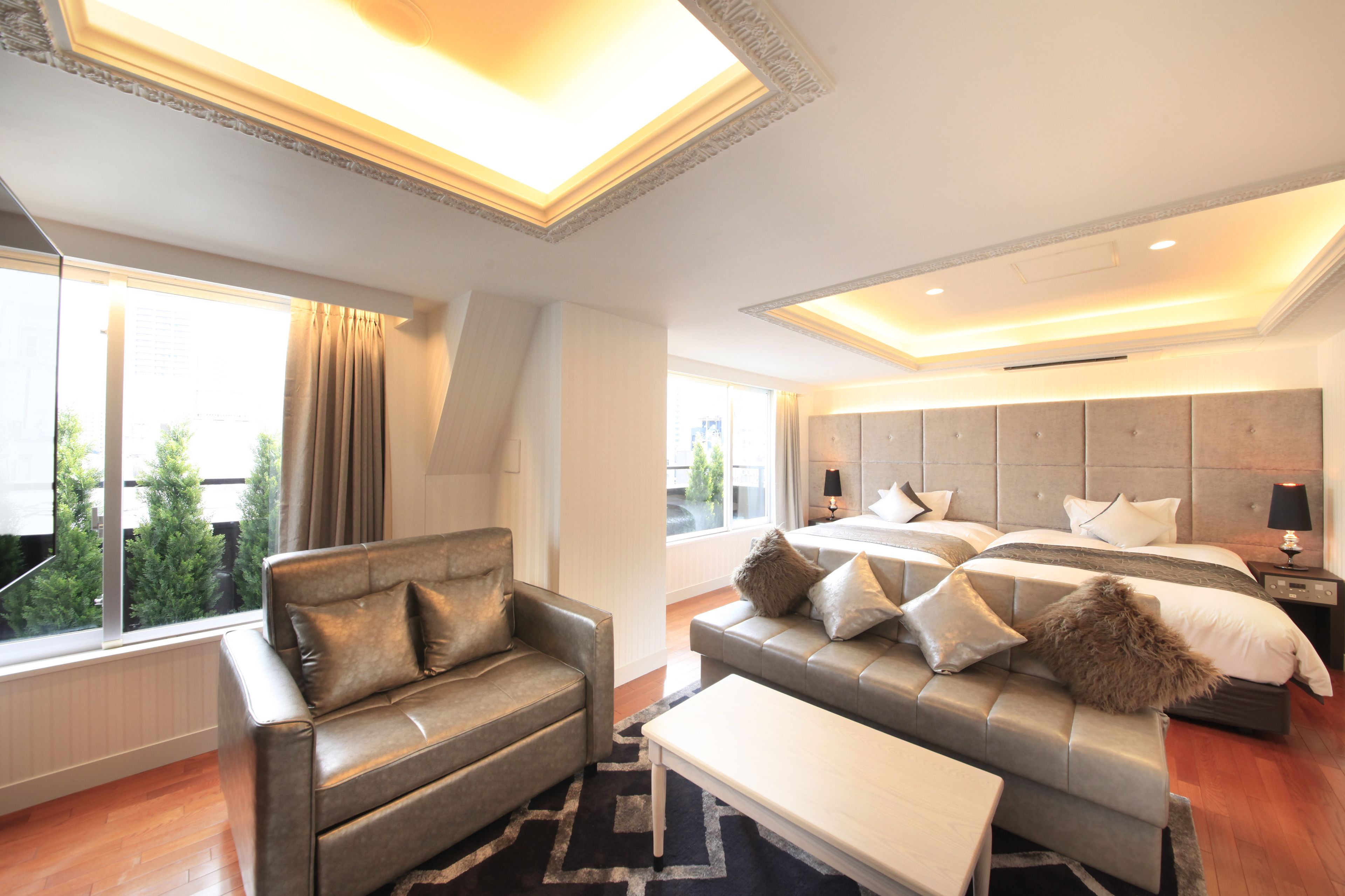 A modern hotel room with a large bed, two sofas, a small table, and a view of greenery outside.