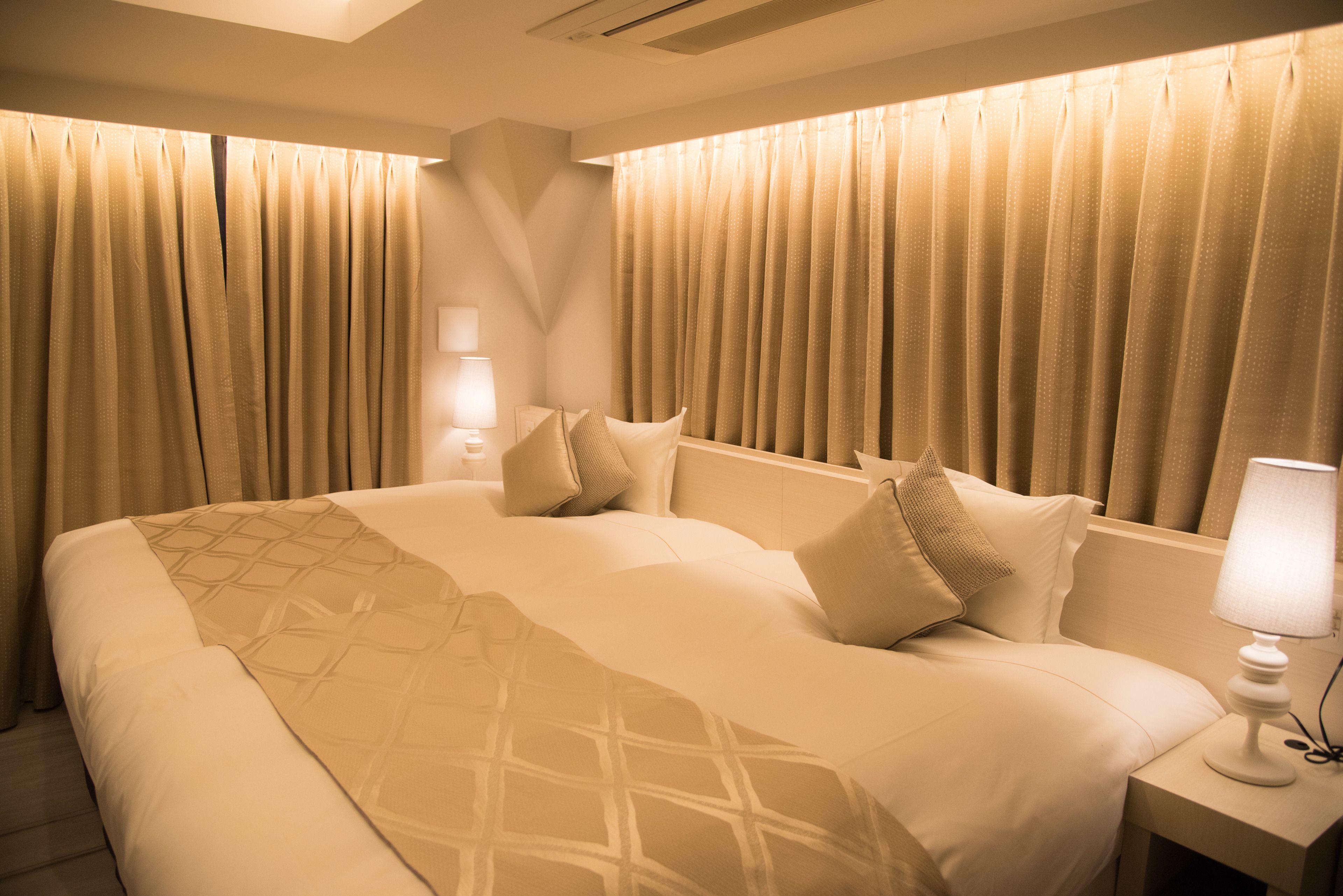 A hotel room with a large bed, beige curtains, a nightstand with a lamp, and a wall-mounted light fixture.
