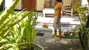 A person standing in a courtyard with a wooden door and a window with a wooden shutter. There is a chair and a table in the background. - Arjuna Homestay Ubud (Ubud)