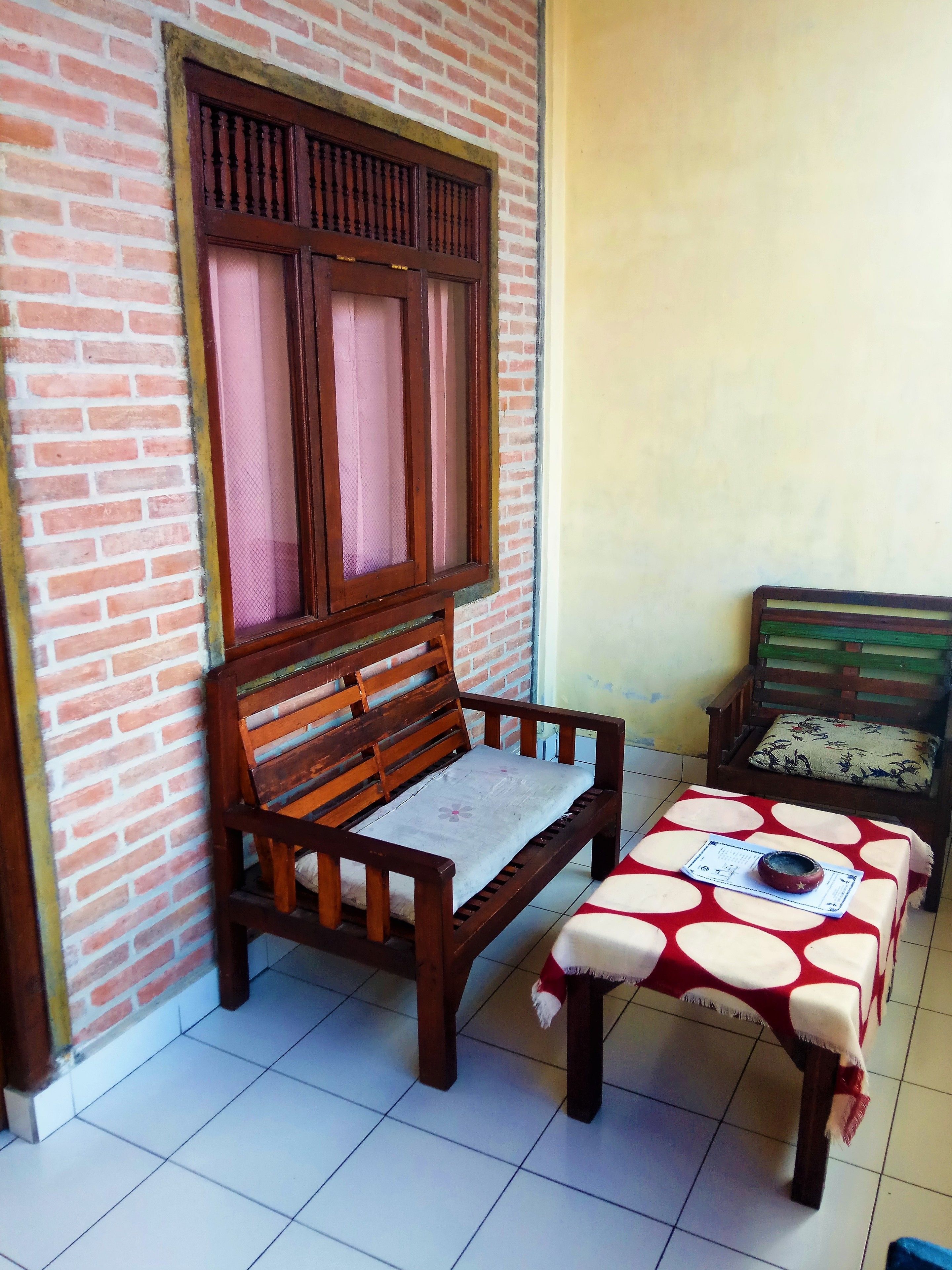 Un banco de madera con cojín, una mesita con mantel de lunares y una ventana con marco de madera y mosquitera.