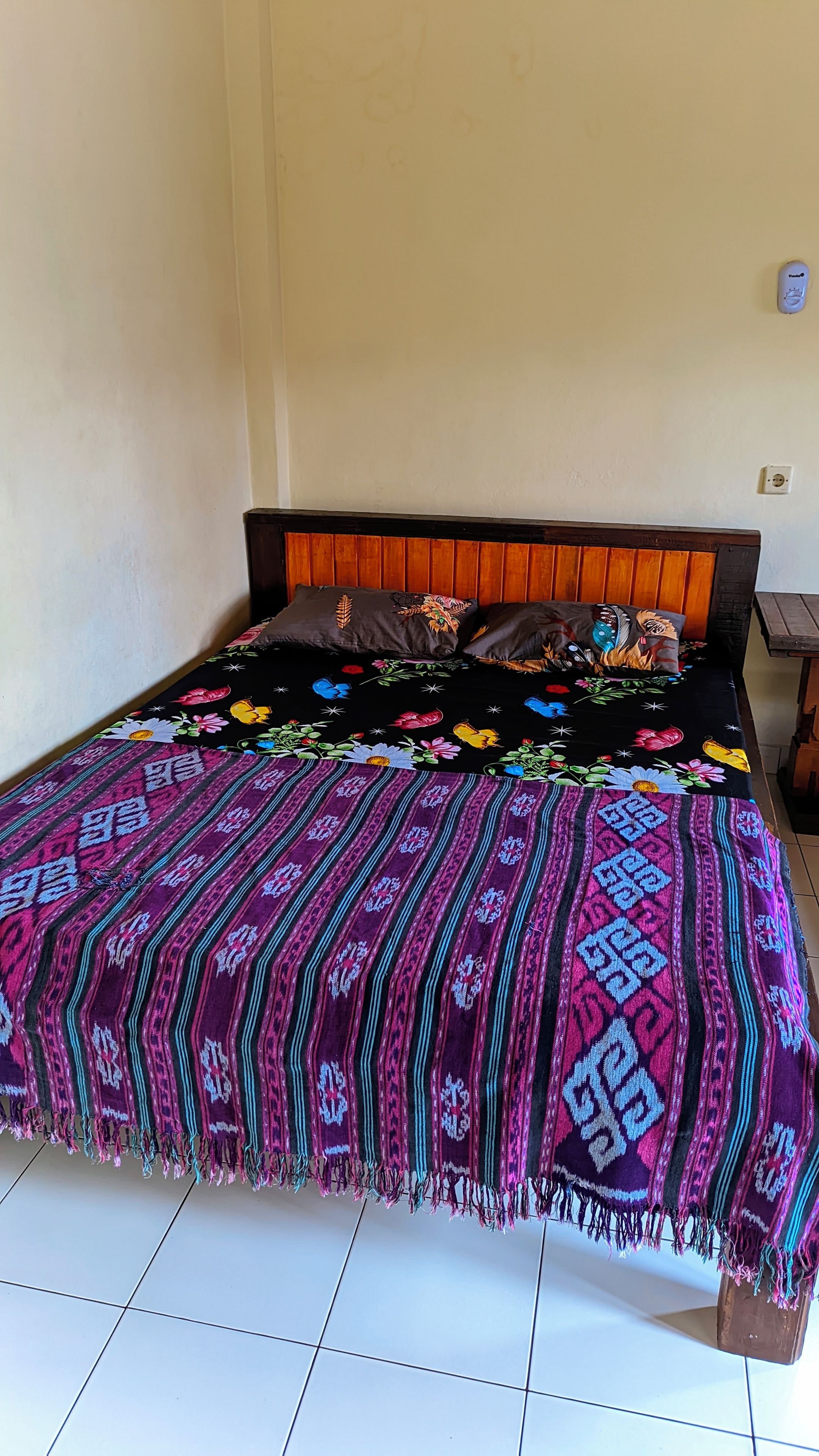 Una cama con una colcha estampada y almohadas, cabecero de madera y suelo de baldosas.