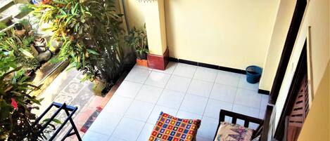 Un balcón con suelo de baldosas, una mesa pequeña con mantel colorido, un banco y varias macetas con plantas.