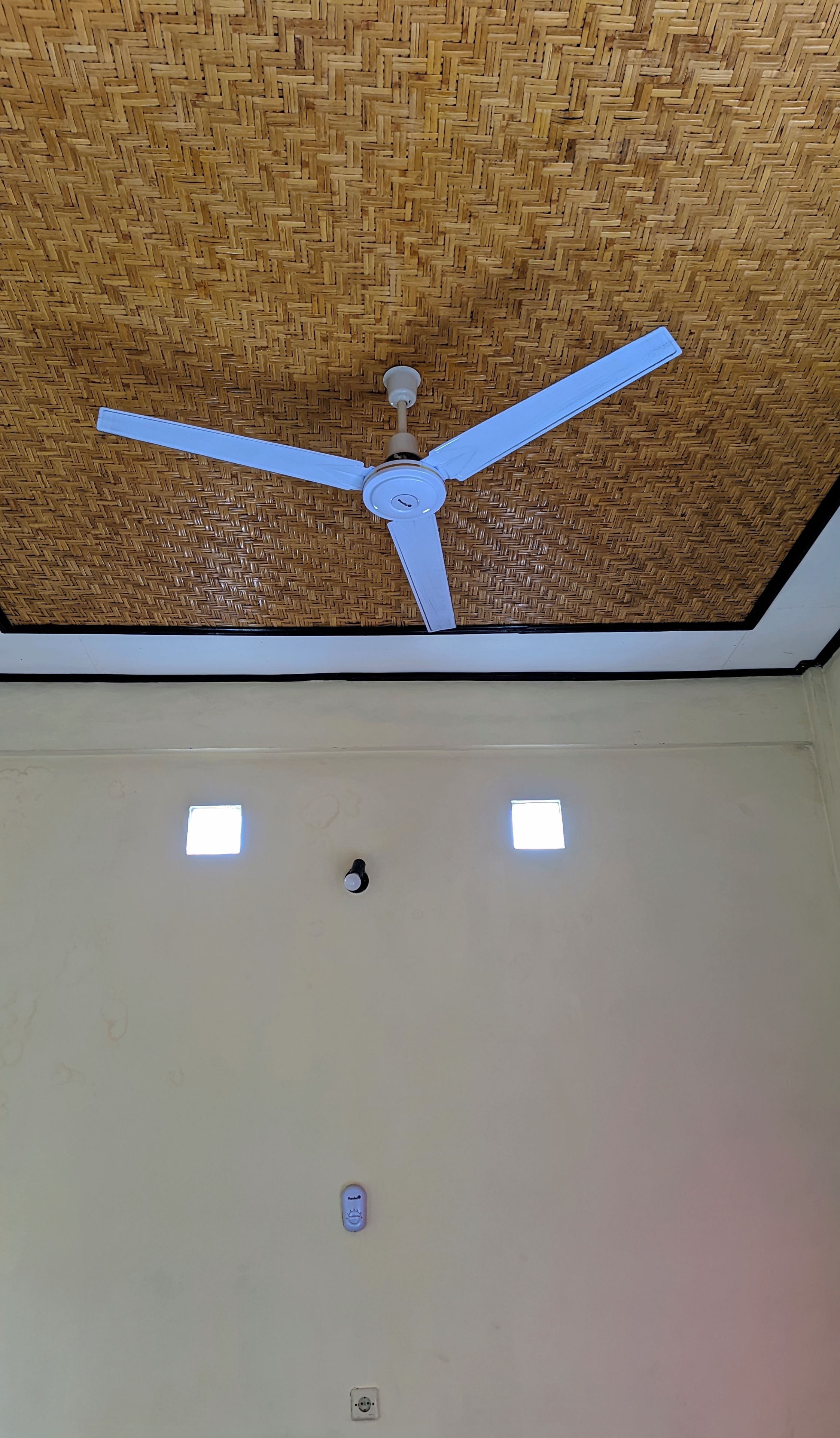 A ceiling fan with three blades, a woven ceiling, and two small windows.