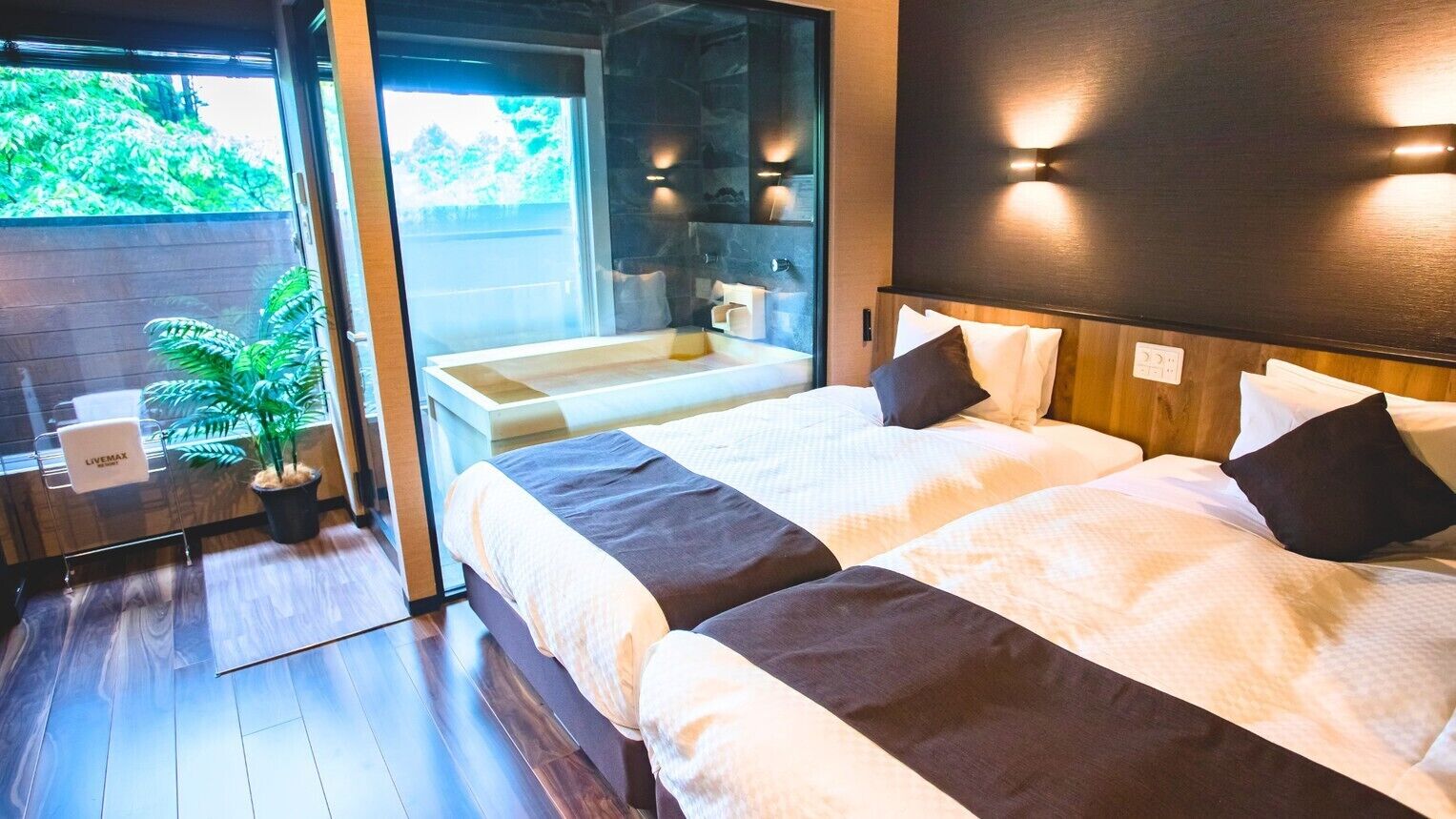 A modern hotel room with two beds, a bathroom with a sink, and a view of greenery outside.