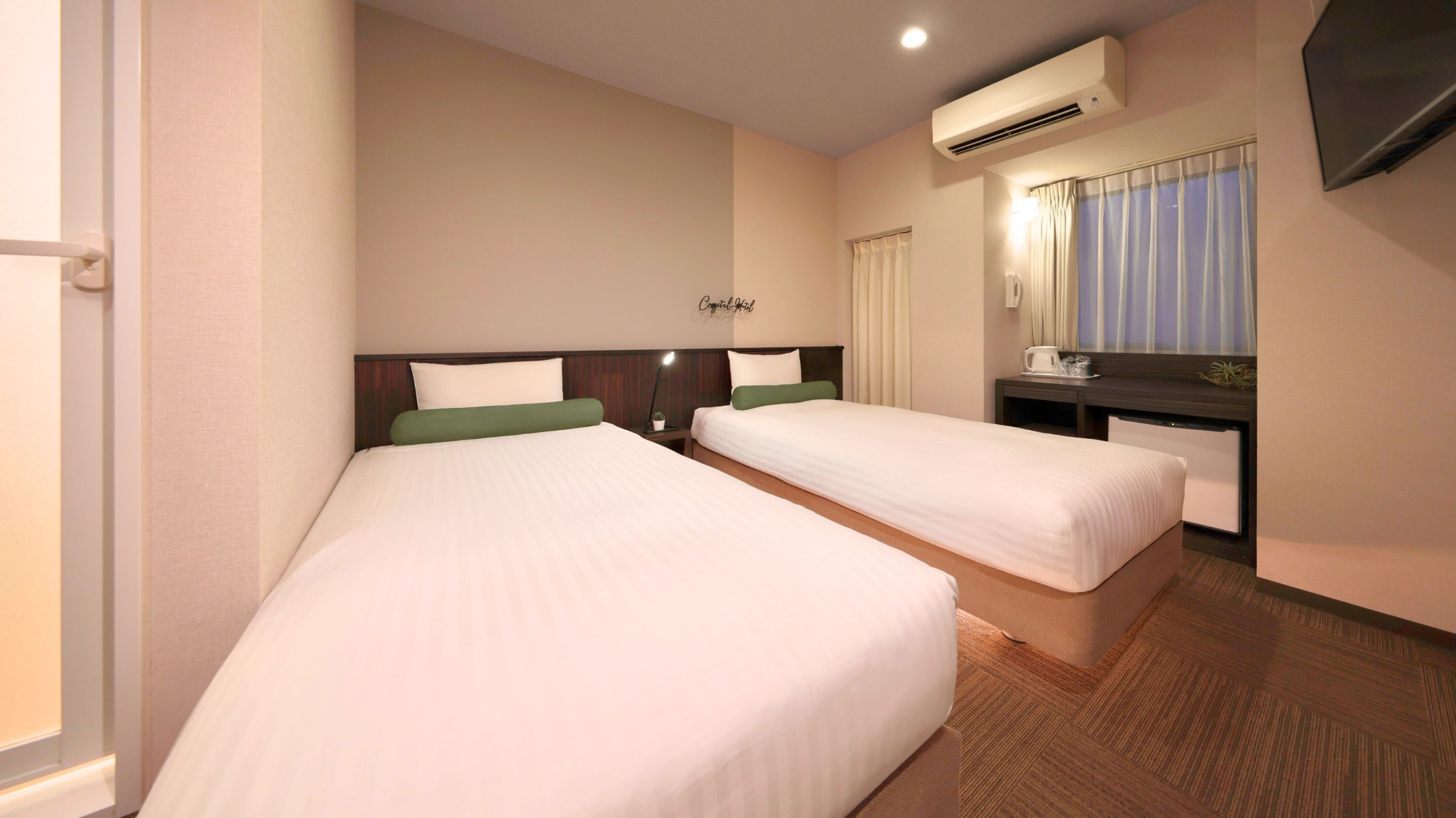 A hotel room with two beds, a desk, a chair, an air conditioning unit, and a window with curtains.