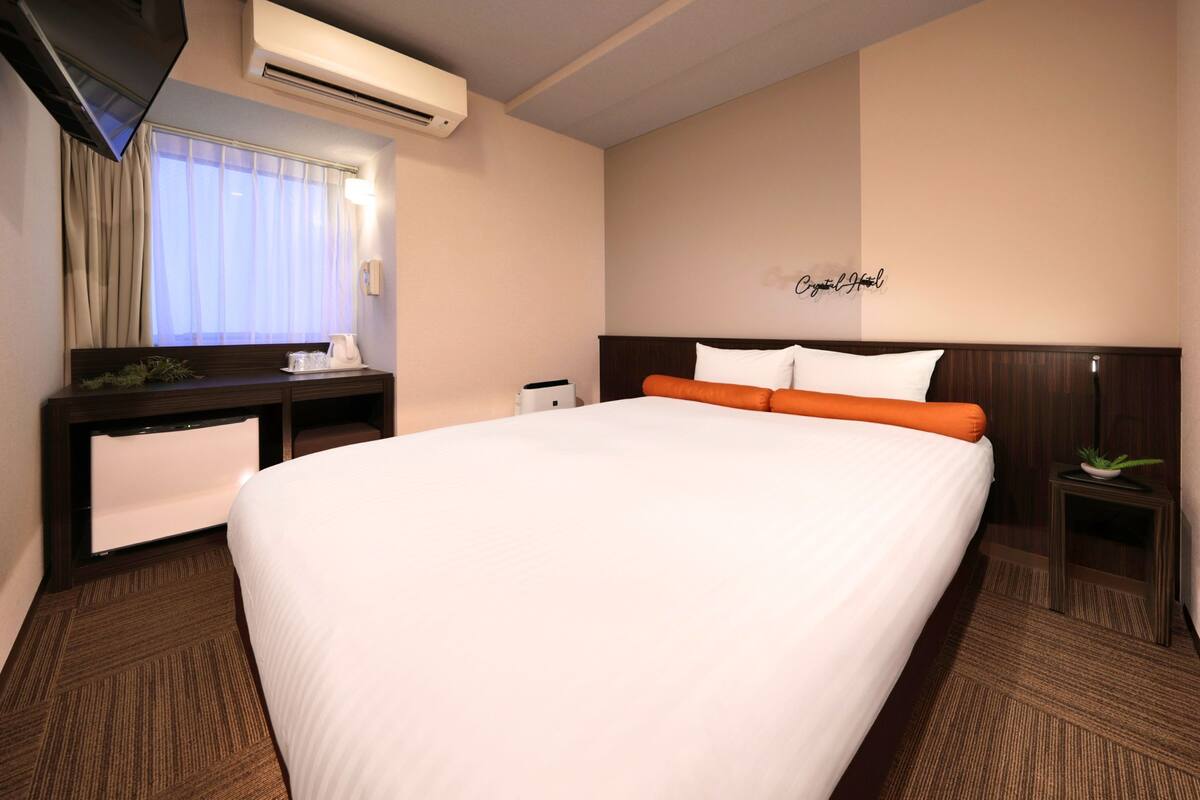 A hotel room with a large bed, a desk with a chair, a TV, and a wall-mounted air conditioner.