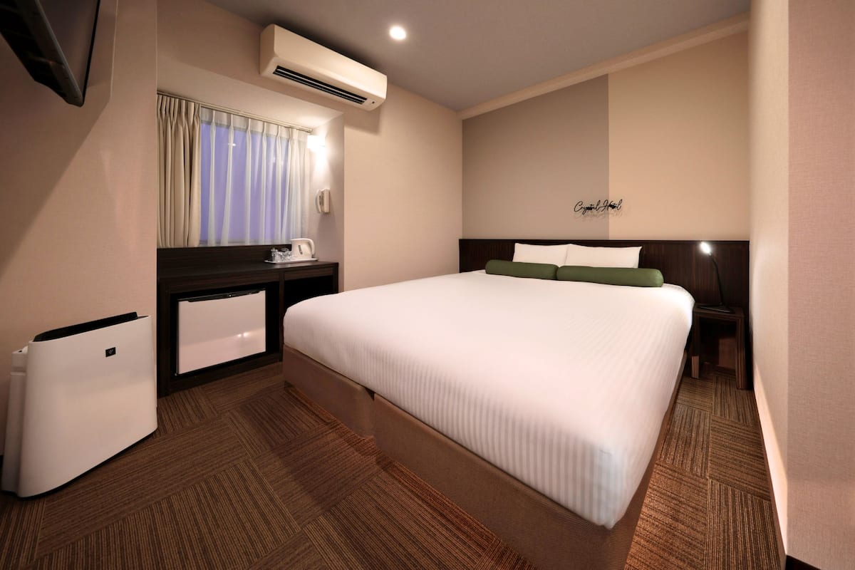 A hotel room with a large bed, a desk with a lamp, a small fridge, and an air conditioning unit.