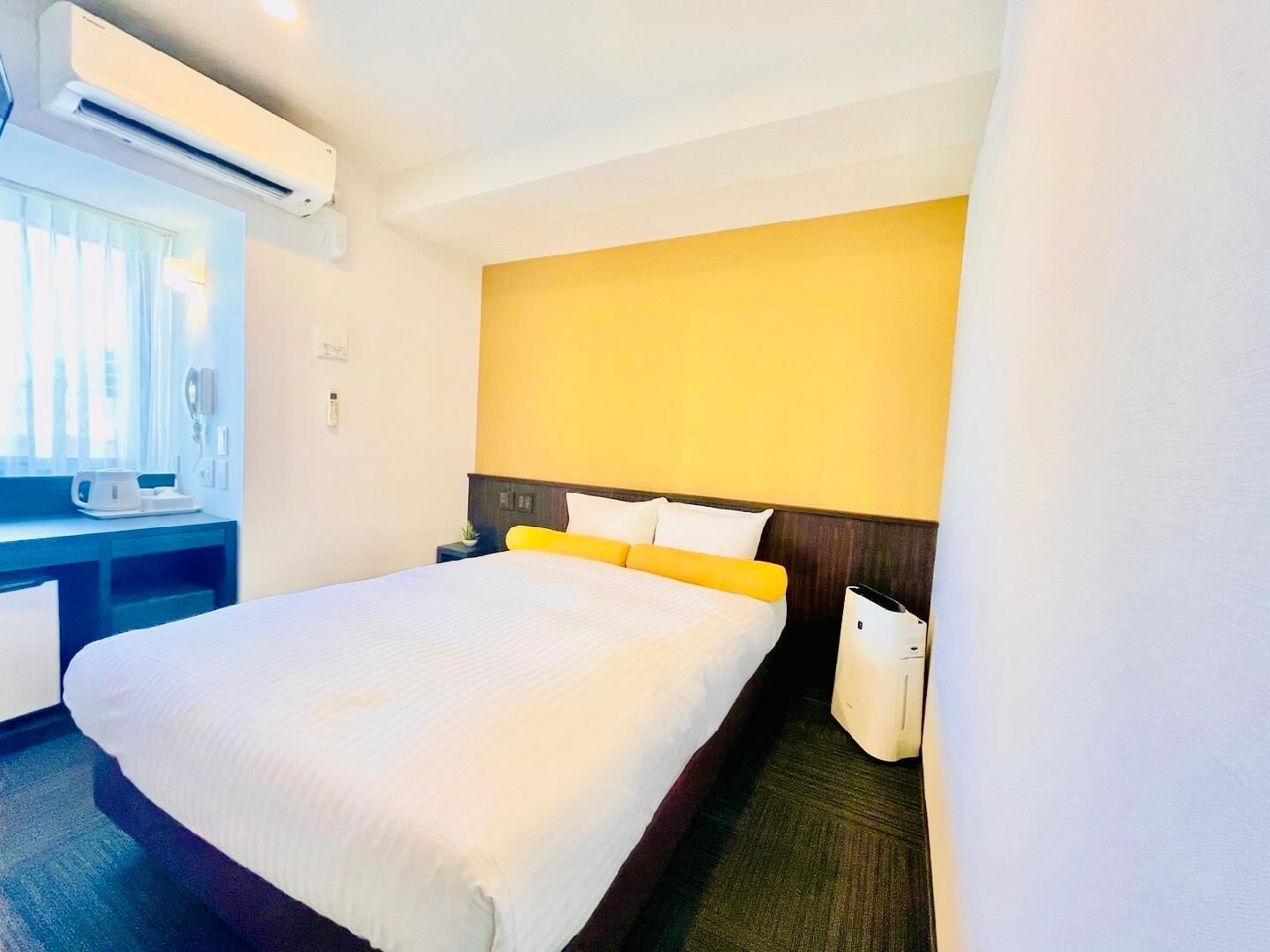 A hotel room with a bed, a desk, an air conditioning unit, and a window.