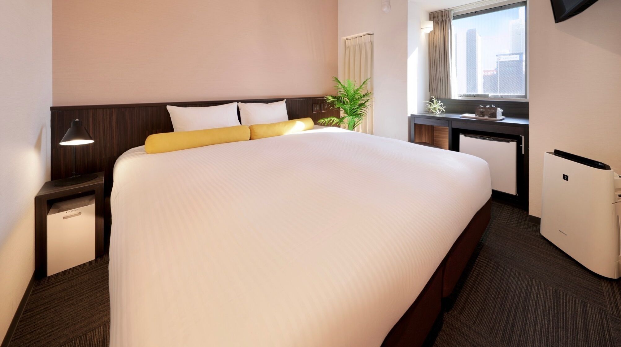 A hotel room with a large bed, bedside tables, a desk, a chair, a plant, and a window with a view of buildings.