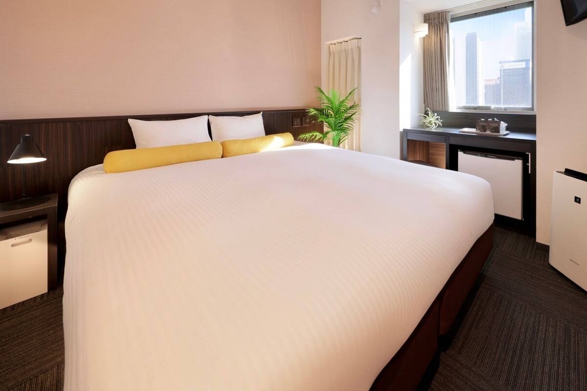 A hotel room with a large bed, bedside tables, a desk, a chair, a plant, and a window with a view of buildings.