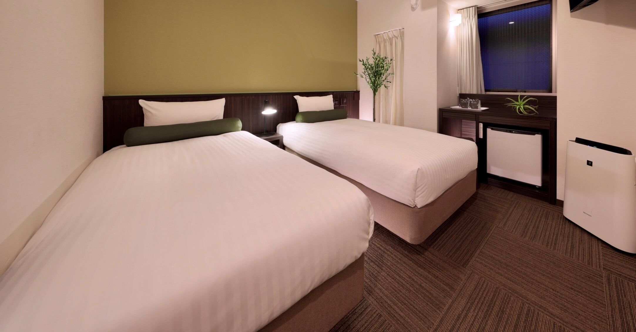 A hotel room with two beds, a desk, a small plant, and a window with curtains.