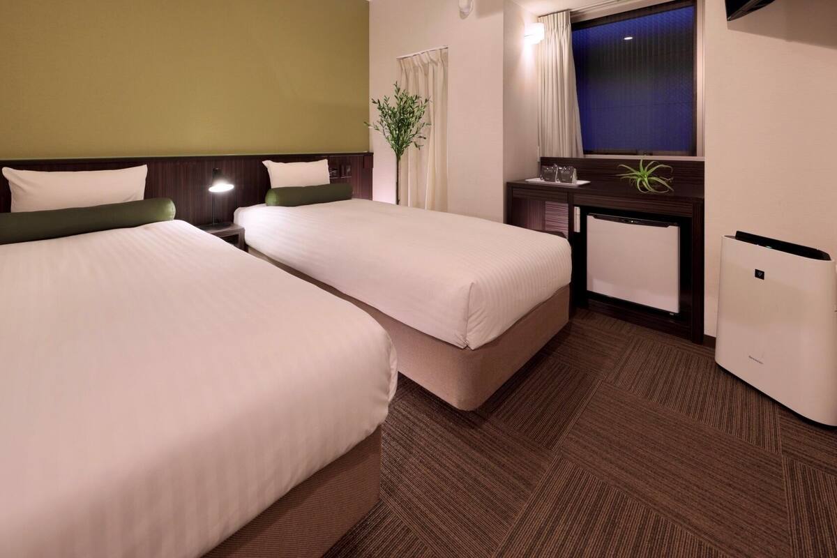 A hotel room with two beds, a desk, a small plant, and a window with curtains.