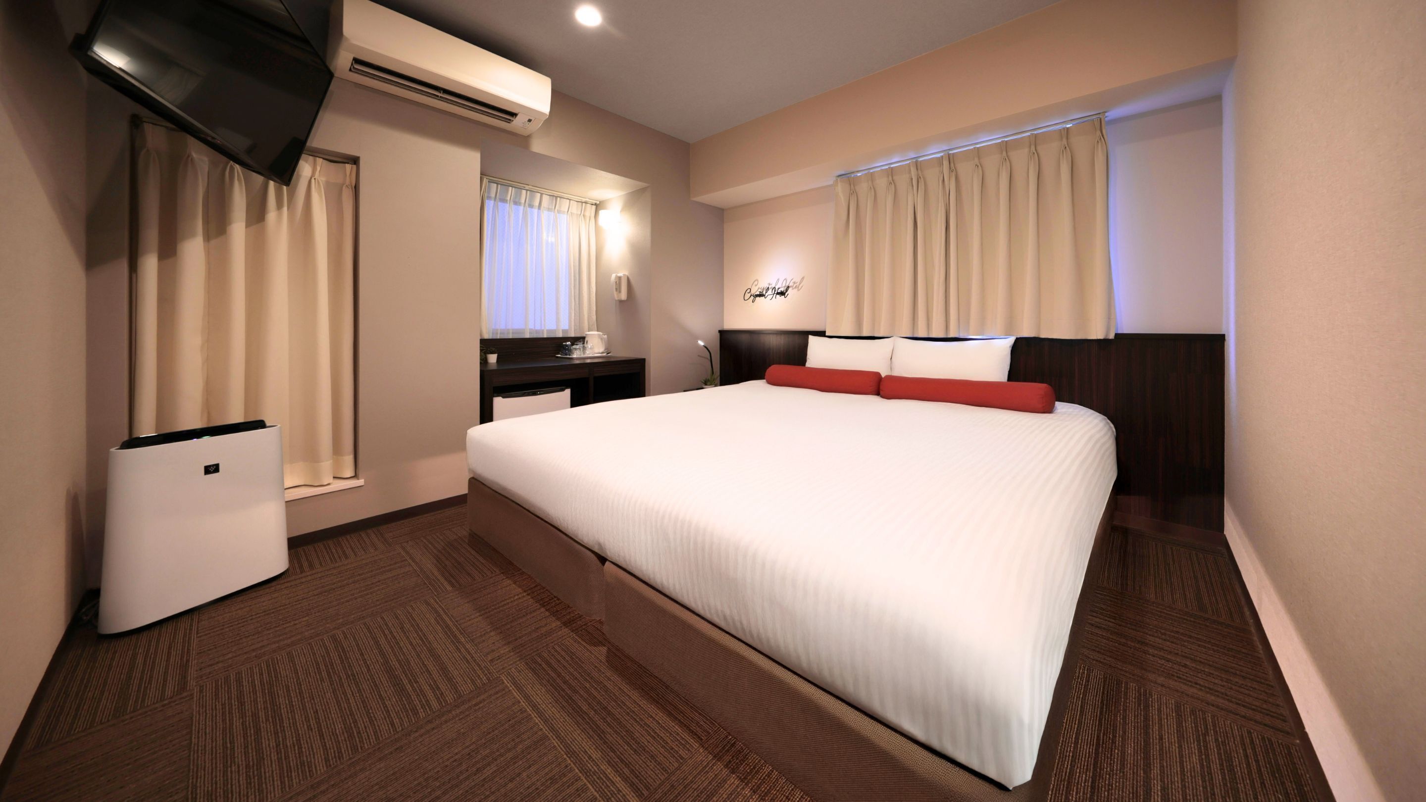 A hotel room with a large bed, a TV, an air conditioning unit, a desk with a chair, and a window with curtains.