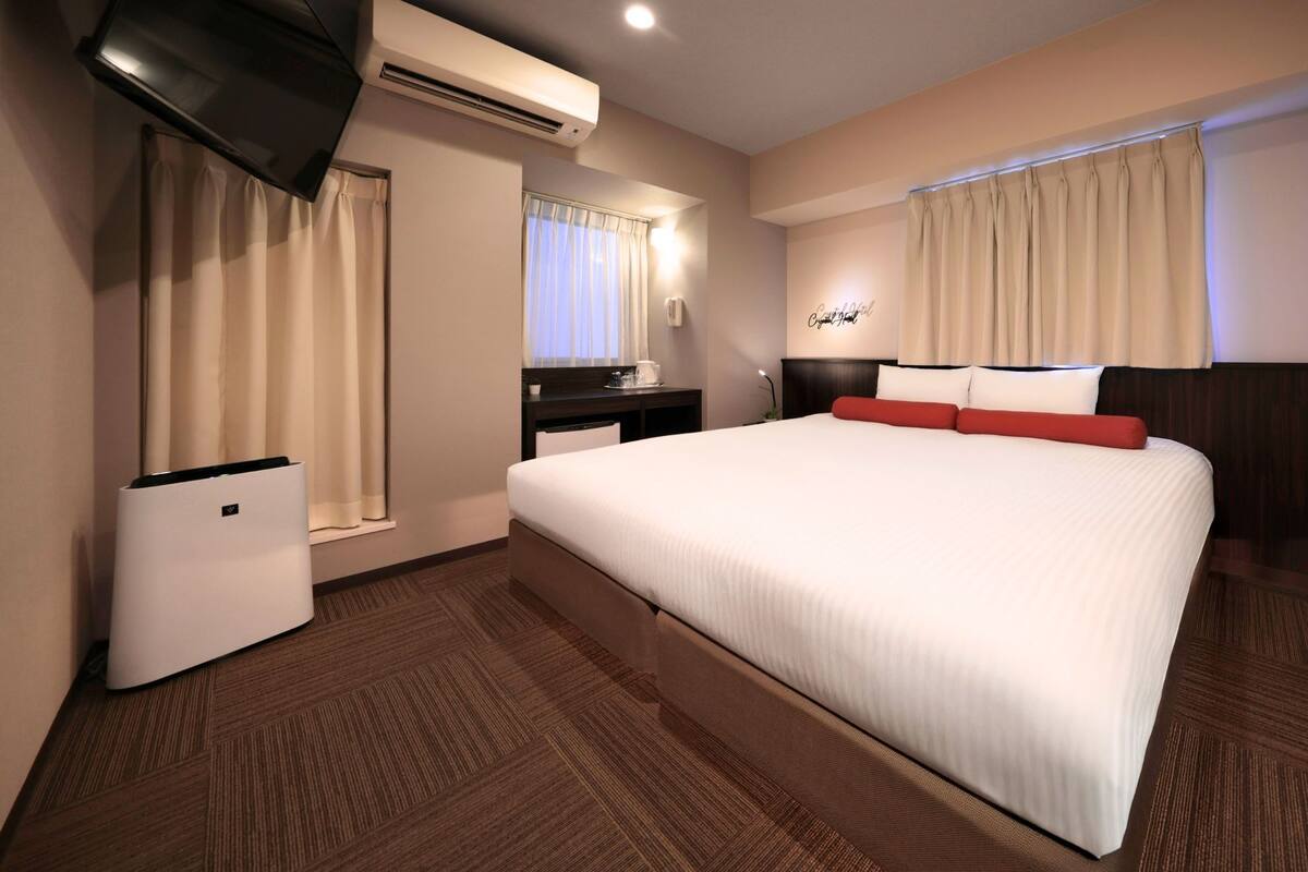 Une chambre d’hôtel avec un grand lit, une télévision, un appareil de climatisation, un bureau avec une chaise et une fenêtre avec des rideaux.