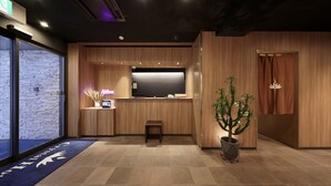 Un área de recepción con un mostrador de madera, un reloj digital, un letrero de neón, una planta pequeña y una alfombra azul con flechas direccionales.
