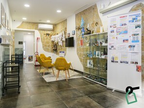 Zona de recepción con pared de piedra, vitrina de vidrio y una pizarra con información. Hay dos sillas amarillas y un escritorio negro con una computadora.