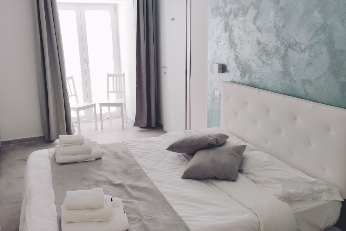 A hotel room with a large bed, grey curtains, a view of the outdoors, and a textured wall.