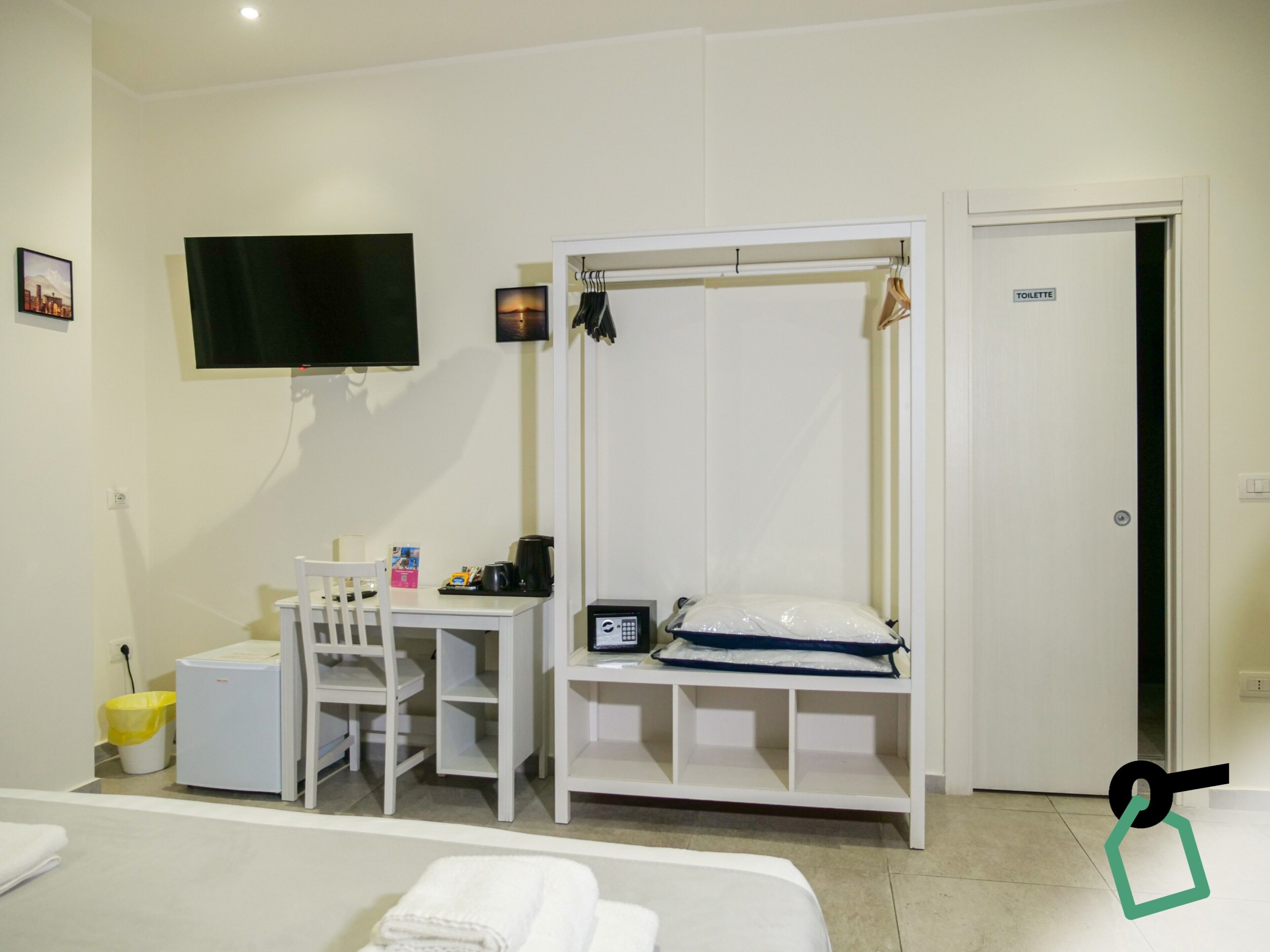 A hotel room with a bed, a desk with a chair, a television, a wardrobe, and a sign indicating a toilet.