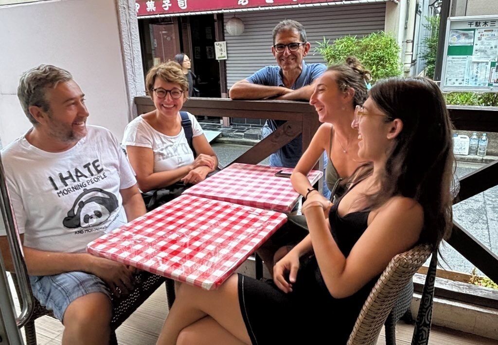 A group of people seated at a table with a red and white checkered tablecloth, outside a building with a sign in a foreign language.