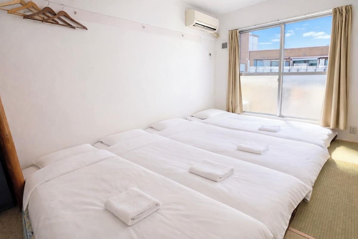 A hotel room with four single beds, a wooden hanger rack, an air conditioning unit, and a large window with curtains.