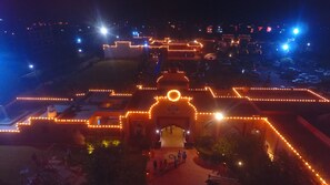 A building with a central dome and surrounding walls illuminated by string lights, with a paved entrance area and a parking lot in the background. - Sadda Pind (Amritsar)