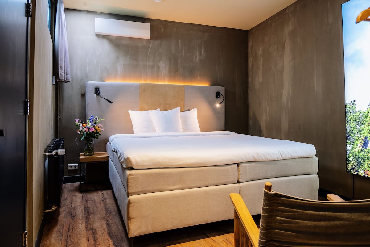 A modern hotel room with a large bed, a bedside table with a lamp, a small desk, and a chair.