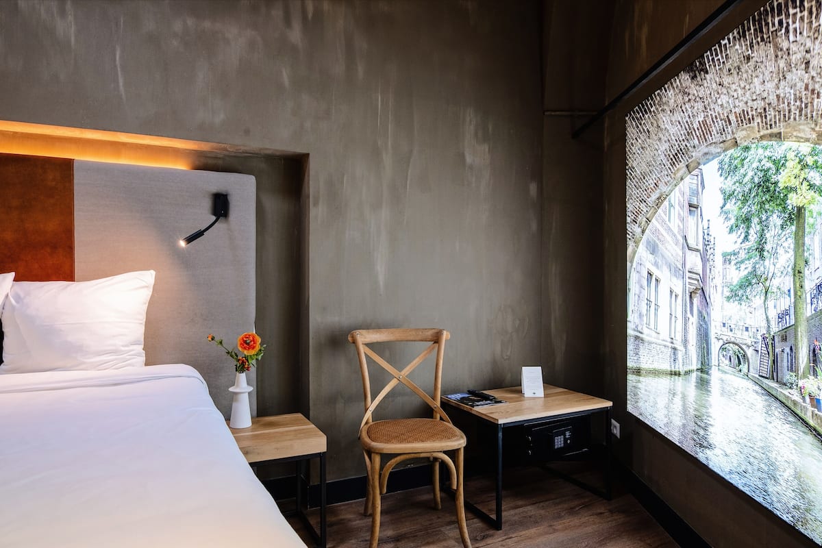 A hotel room with a large bed, a wooden chair, a small table, and a view of a canal through a window.