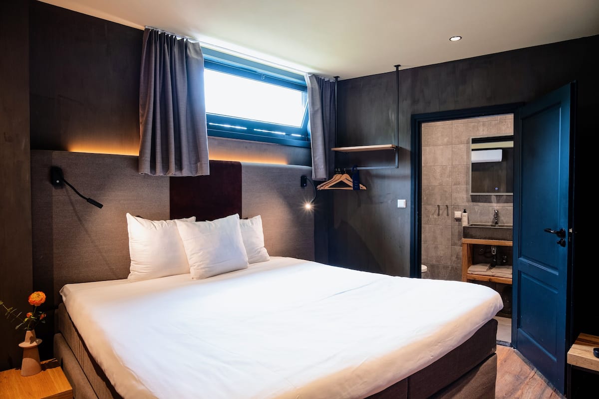 A modern hotel room with a large bed, a bedside table with a vase of flowers, a wall-mounted light, and a view of the bathroom through an open door.