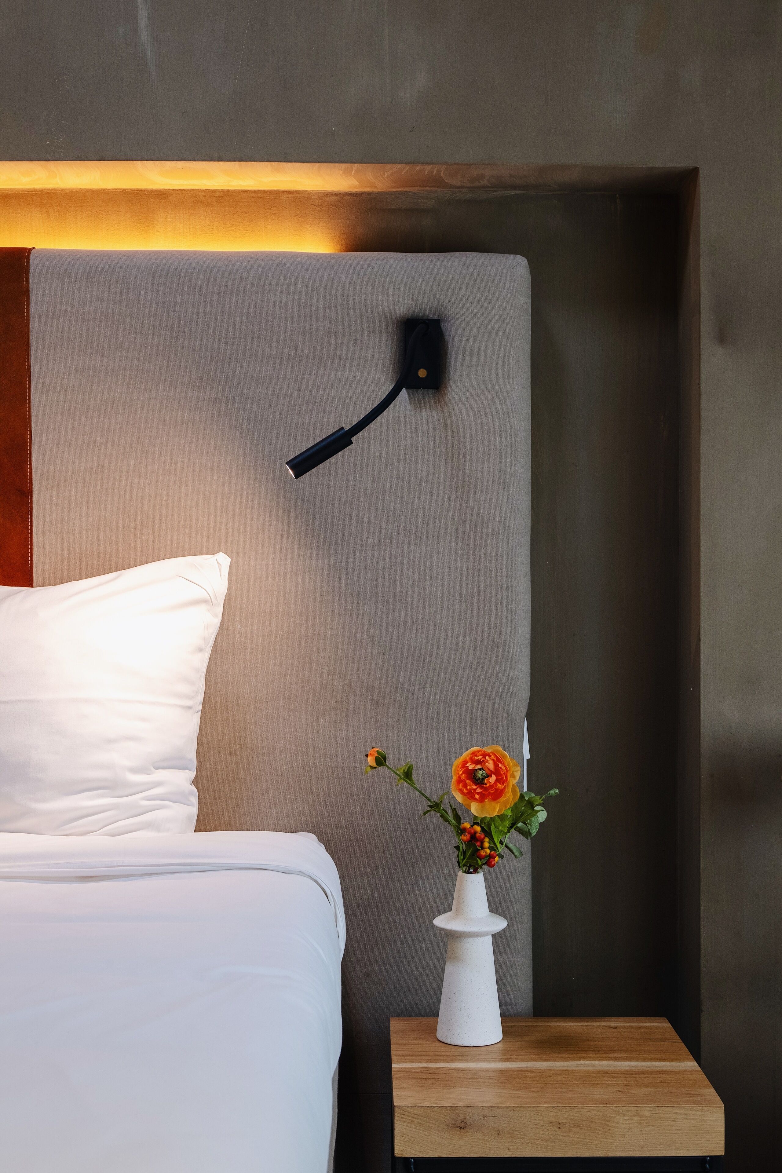 A neatly made bed with white bedding, a bedside table with a white vase containing a single flower, and a wall-mounted light fixture.