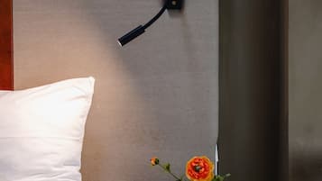 A neatly made bed with white bedding, a bedside table with a white vase containing a single flower, and a wall-mounted light fixture.