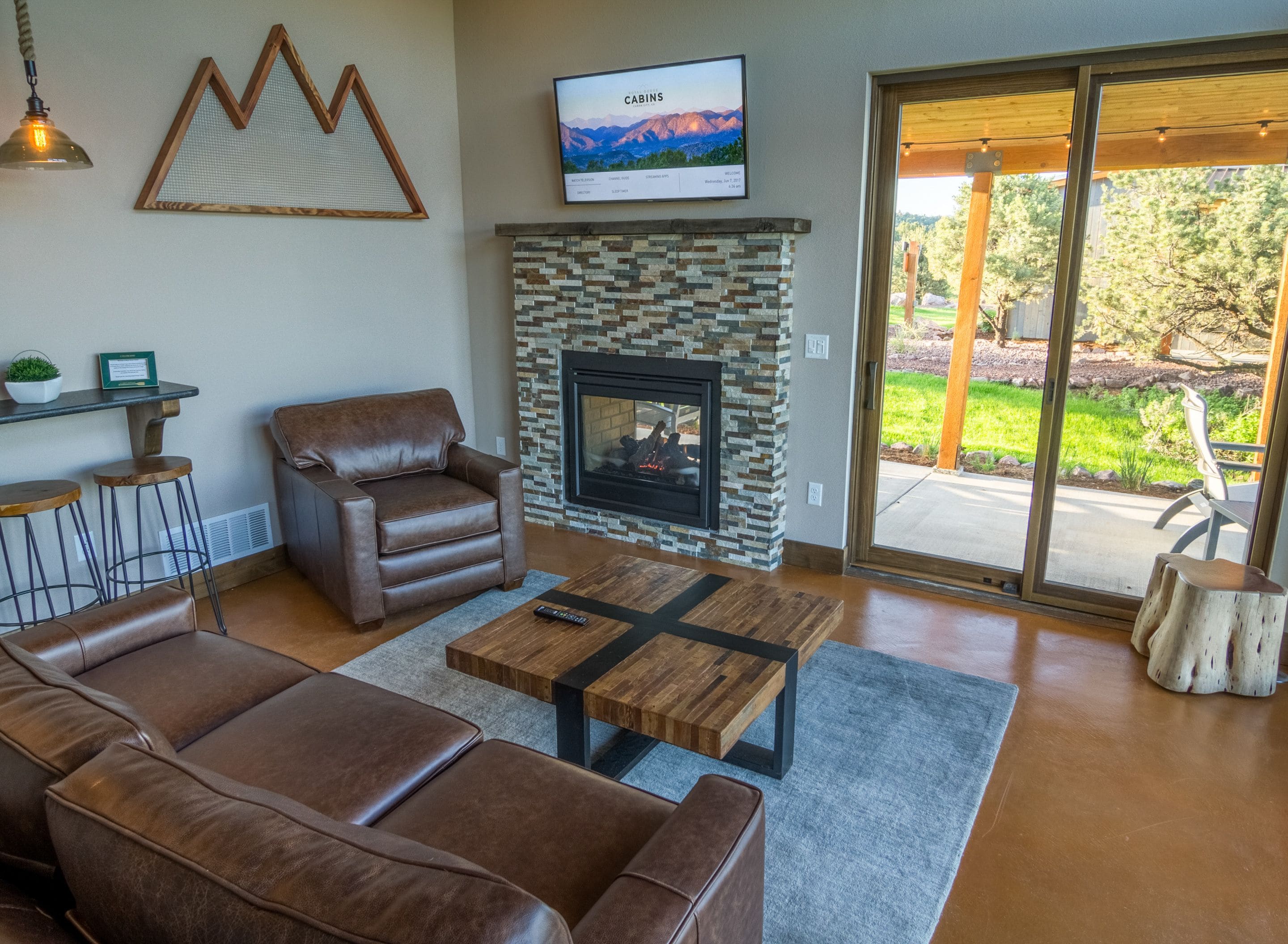 A living room with a brown leather sofa, a brown leather armchair, a wooden coffee table, a stone fireplace, and a television mounted on the wall.