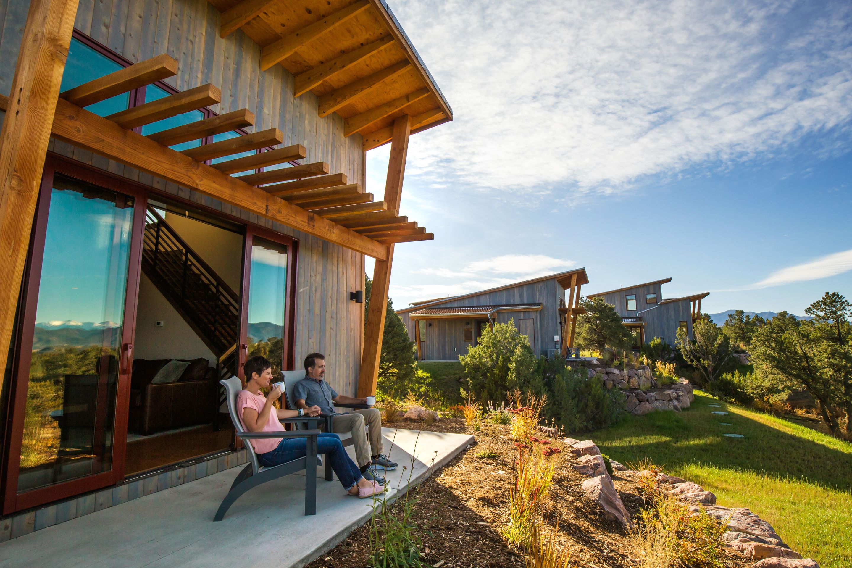 A modern house with a wooden exterior, large glass windows, and a patio area where two people are seated.