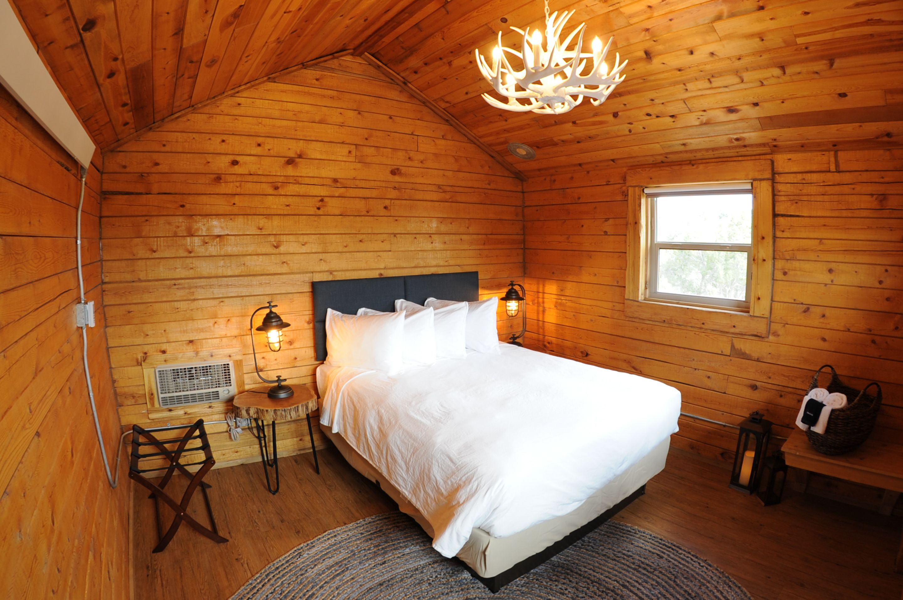 Camera da letto in una baita di legno con un letto, due lampade a parete, una finestra e un lampadario.