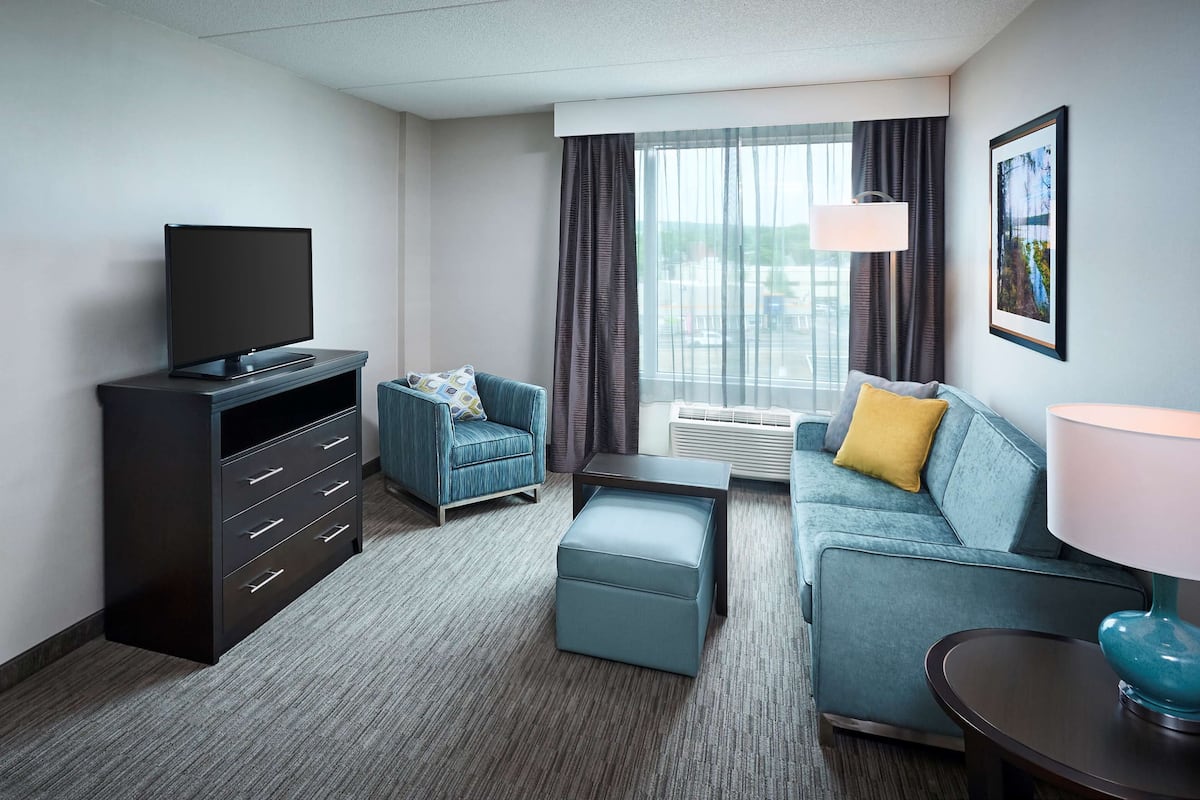 Ein Hotelzimmer mit Bett, Fernseher, blauem Sessel, Sofa, Couchtisch und einem Fenster mit Vorhängen.